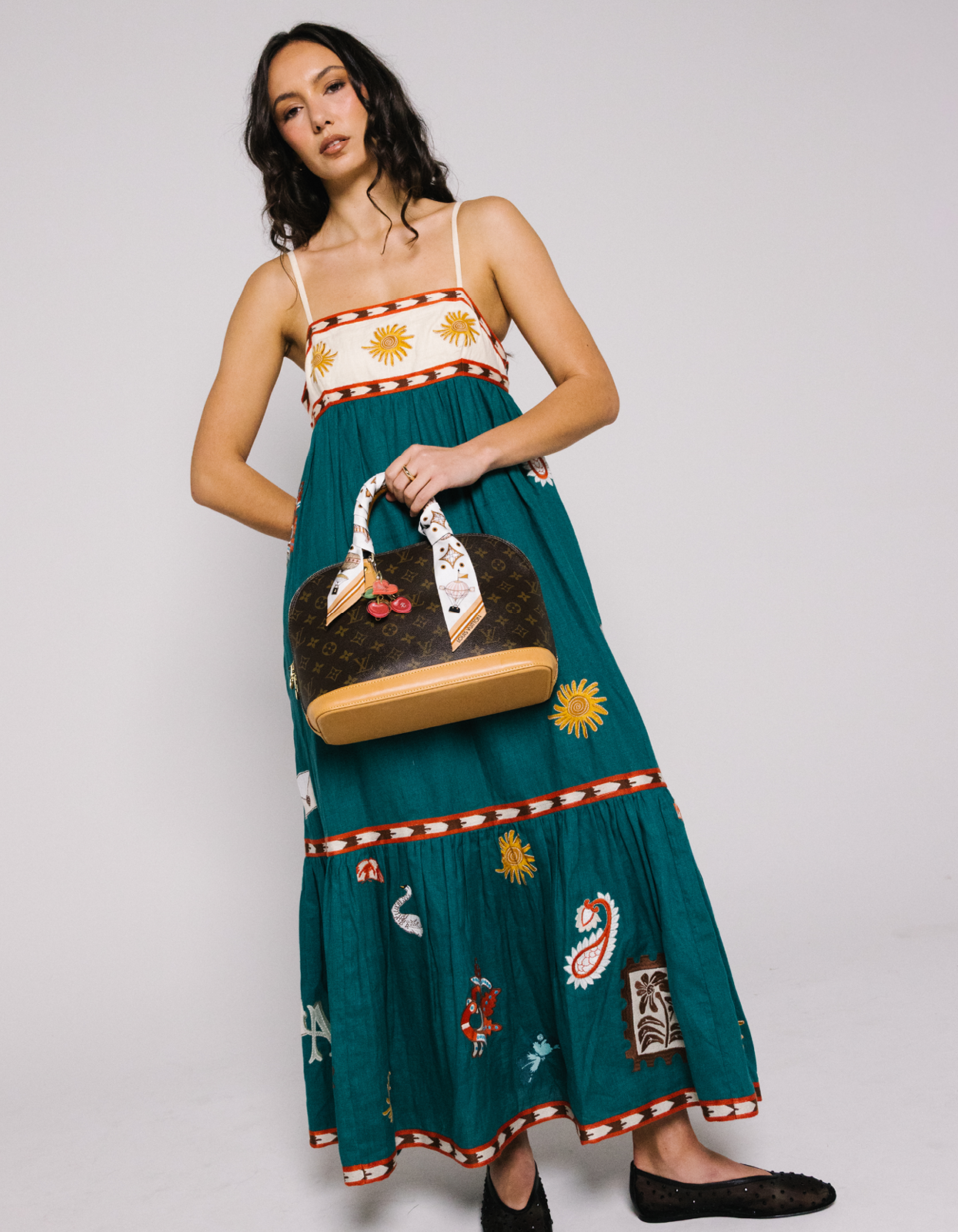 A woman modeling a teal maxi dress with embroidered details, holding a brown monogram handbag against a light gray studio background.